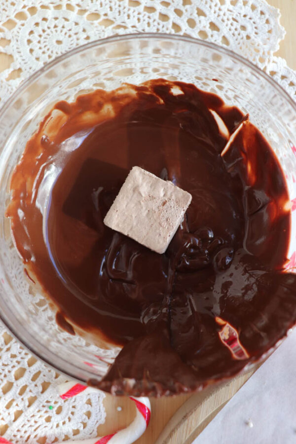 A glass bowl filled with melted chocolate, with a square piece of fudge being dipped in the center. The bowl sits on a lacy white doily atop a wooden surface.