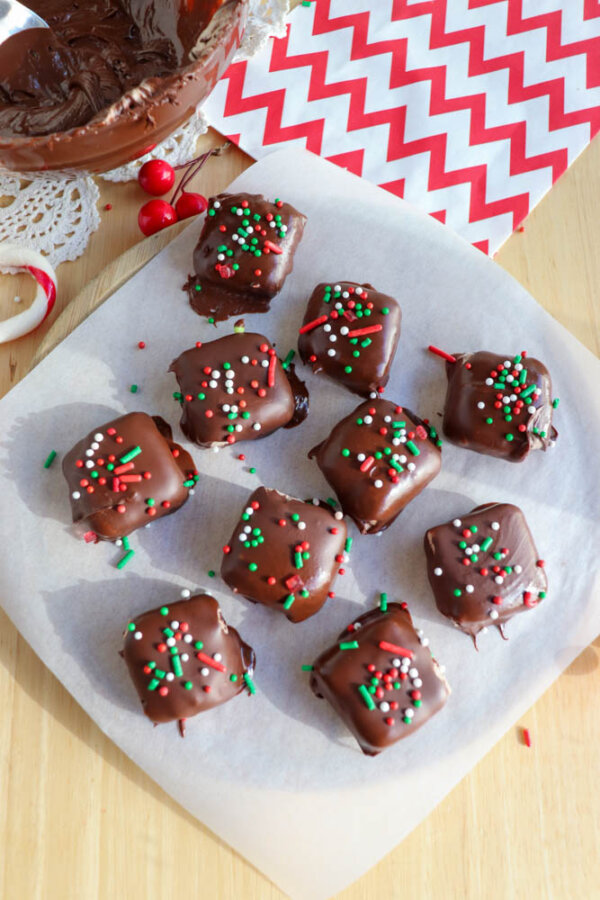Nine chocolate-covered square treats with red, white, and green sprinkles on parchment paper. A bowl of melted chocolate, a spoon, and red-and-white chevron napkins are nearby. The scene is festive and bright.