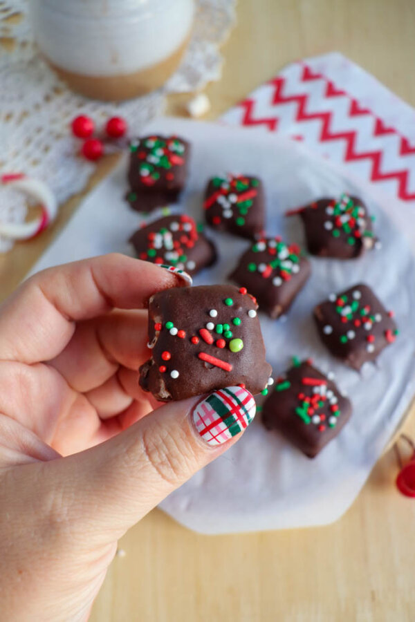 A hand holding a chocolate-covered treat decorated with red, green, and white sprinkles. More similarly decorated treats are on parchment paper in the background, with festive napkins and a lace cloth nearby.