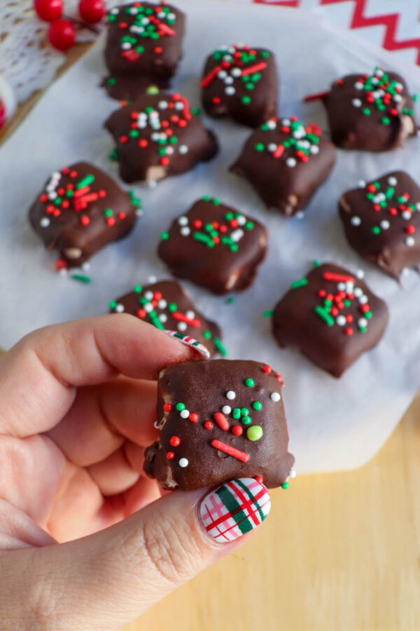 A hand with festive plaid nail art holds a chocolate-covered treat topped with red, green, and white sprinkles. More similar treats are arranged on parchment paper in the background.