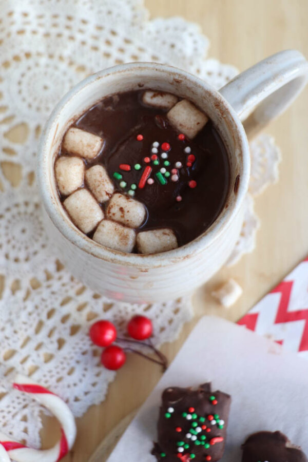 A mug of hot chocolate topped with mini marshmallows and red, green, and white sprinkles sits on a lace doily, with festive decorations and chocolate treats nearby.