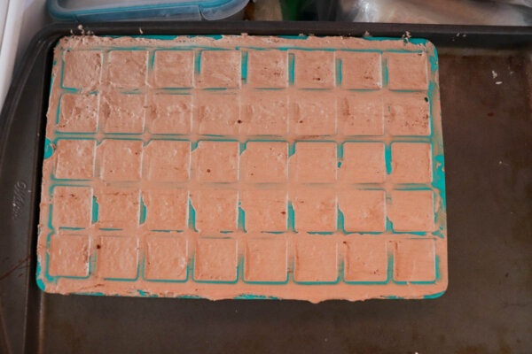 A blue silicone ice cube tray filled with a light brown mixture sits on a baking sheet, likely in preparation for freezing.