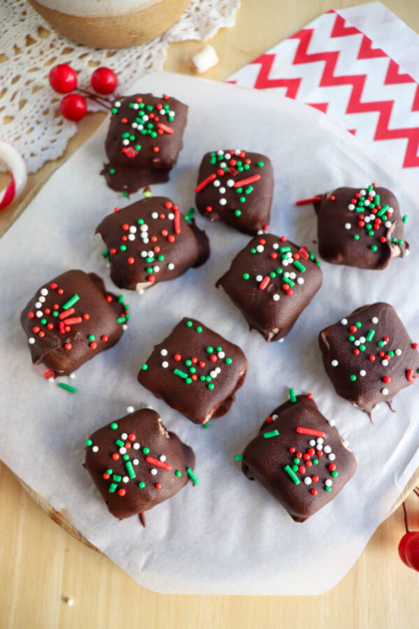 Nine square chocolate-covered treats topped with red, green, and white sprinkles are arranged on parchment paper. Some cranberries and a red-and-white napkin are in the background.