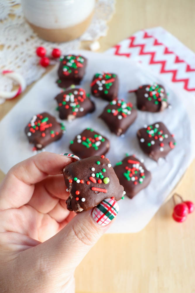 A hand holding a chocolate-covered treat with festive red, green, and white sprinkles. More decorated treats are on a piece of parchment paper in the background, with a red and white napkin nearby.