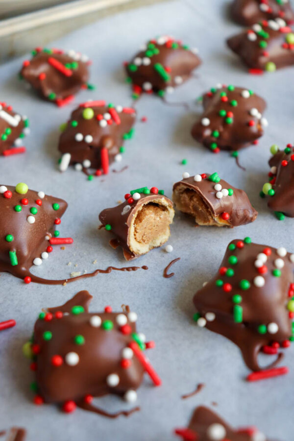 Bite-sized chocolate-covered treats with colorful red, green, and white sprinkles are arranged on parchment paper. One treat is cut open, revealing a creamy peanut butter filling inside.