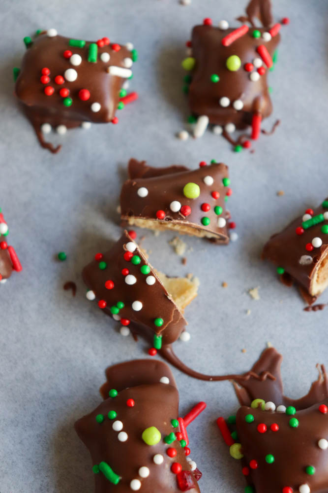 Squares of chocolate-covered treats topped with red, white, and green sprinkles are scattered on a surface, with a few pieces broken open to reveal a light, flaky interior.
