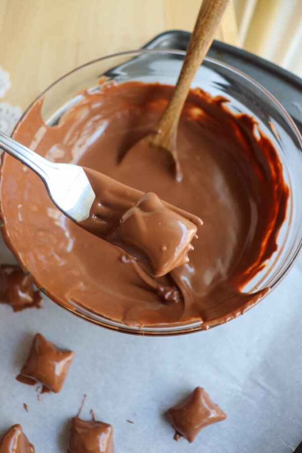 A fork holds a square candy coated in melted chocolate over a glass bowl of more melted chocolate, with other chocolate-coated candies resting on parchment paper nearby. A wooden spoon is in the bowl.