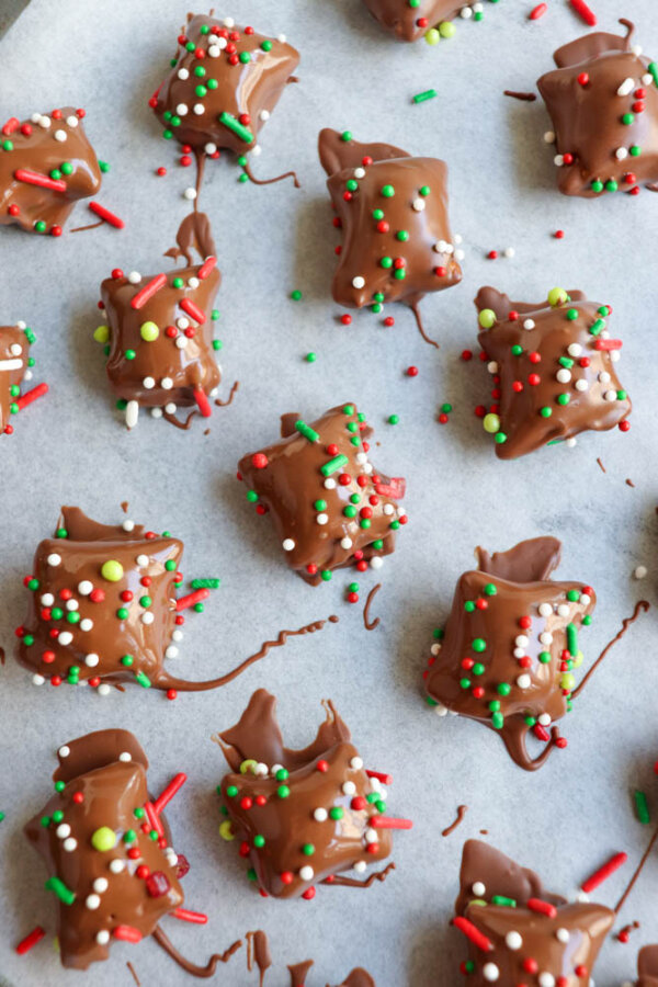Chocolate-covered treats topped with colorful red, green, and white sprinkles are arranged on a sheet of parchment paper. The treats appear to be cooling or setting, with some chocolate and sprinkles scattered around.