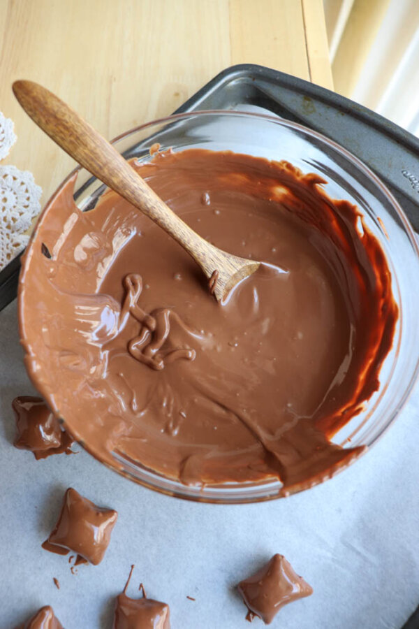 A glass bowl filled with melted milk chocolate and a wooden spoon rests on a metal tray, with several chocolate-coated candies on parchment paper beside it.