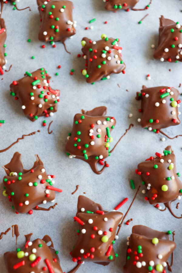 Chocolate-covered treats topped with red, green, and white sprinkles are scattered on a sheet of parchment paper. Some chocolate drizzles are visible around the candies.