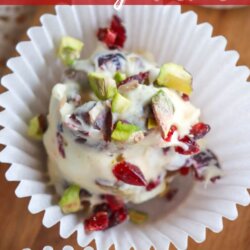White chocolate clusters with chopped pistachios and dried cranberries are displayed in white paper cups on a wooden surface. Text reads: “Crockpot pistachio & cranberry clusters.”.