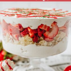 A glass trifle bowl filled with layers of cake, whipped cream, and sliced strawberries sits on a table with Christmas decorations. Text reads: โLittle Debbie Christmas tree cake trifleโ and โhomemadeheather.com.โ.
