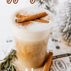 A creamy eggnog latte topped with cinnamon sticks and sprinkled with cinnamon sits on a table with rosemary sprigs, holiday decorations, and text reading eggnog latte and homemade above and below the drink.
