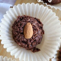 A close-up of homemade chocolate coconut almond treats on white cupcake liners, each topped with a whole almond, displayed on a wooden surface. Text reads Crockpot Almond Joy and homemadeheather.com.
