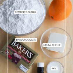 A bowl of powdered sugar, an orange, cream cheese, a small glass of milk, a bottle of orange extract, and a box of dark chocolate are arranged on a wooden surface. Each item is labeled.