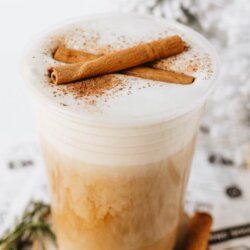 A plastic cup of eggnog latte topped with foam, cinnamon powder, and a cinnamon stick, sits on a wooden coaster with a sprig of rosemary beside it. The text reads EGGNOG latte and HOMEMADEHEATHER.COM.