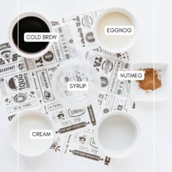Flat lay of ingredients in bowls labeled cold brew, eggnog, cream, syrup, and nutmeg, arranged on a white surface with patterned paper. Website homemadeheather.com is at the bottom.