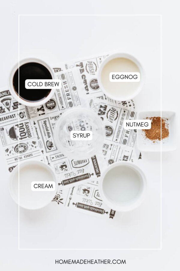 Flat lay of ingredients in bowls labeled cold brew, eggnog, cream, syrup, and nutmeg, arranged on a white surface with patterned paper. Website homemadeheather.com is at the bottom.