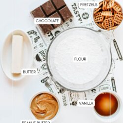 A flat lay of baking ingredients in bowls labeled: pretzels, chocolate, flour, butter, vanilla, and peanut butter, arranged on a white surface with a decorative paper underneath.