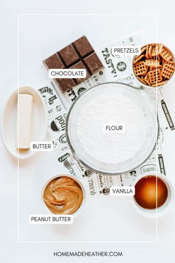 A flat lay of baking ingredients in bowls labeled: pretzels, chocolate, flour, butter, vanilla, and peanut butter, arranged on a white surface with a decorative paper underneath.