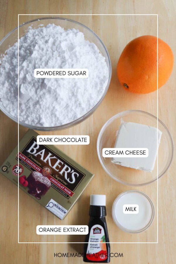 A bowl of powdered sugar, an orange, cream cheese, a small glass of milk, a bottle of orange extract, and a box of dark chocolate are arranged on a wooden surface. Each item is labeled.