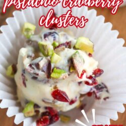 A white chocolate cluster with chopped pistachios and cranberries sits in a white paper cup on a wooden surface. Text reads: “white chocolate pistachio cranberry clusters” and “CRAZY GOOD!”.