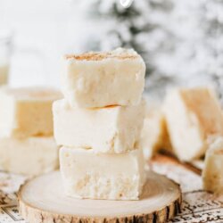 Three pieces of creamy eggnog fudge are stacked on a wooden slice, with more fudge pieces in the background. Text reads Eggnog fudge at the top and homemadeheather.com at the bottom.
