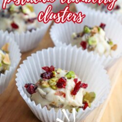 White chocolate pistachio cranberry clusters in white paper cups, topped with chopped pistachios and dried cranberries. Text overlay reads, White Chocolate Pistachio Cranberry Clusters - Crockpot Recipe.