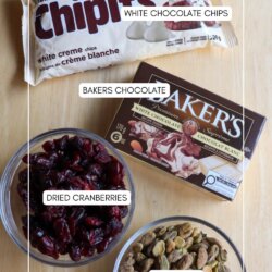A flat lay of four ingredients on a wooden surface: a bag of white chocolate chips, a box of Bakers white chocolate, a bowl of dried cranberries, and a bowl of shelled pistachios. Each item is labeled.