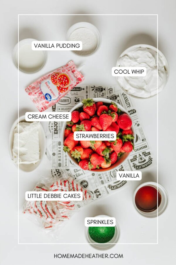 A flat lay of ingredients for a dessert: a bowl of strawberries, vanilla pudding, cool whip, cream cheese, vanilla, Little Debbie cakes, and containers of red and green sprinkles are all labeled and arranged on a white surface.