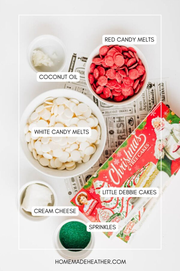 A flat lay of baking ingredients: bowls of white and red candy melts, coconut oil, cream cheese, green sprinkles, and a box of Little Debbie Christmas Tree Cakes on vintage papers—each item is labeled.
