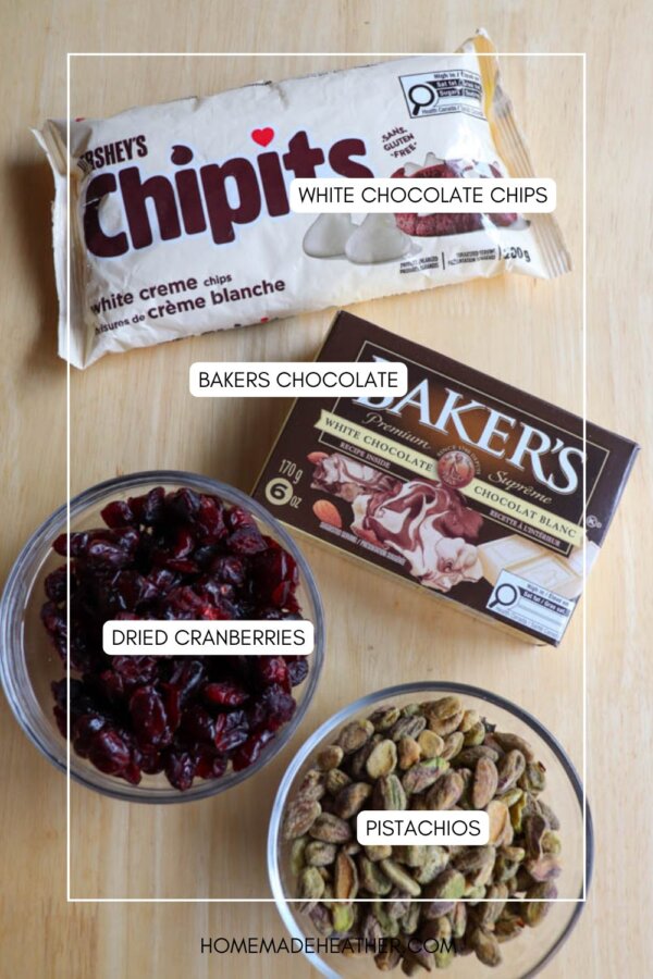 A flat lay of four ingredients on a wooden surface: a bag of white chocolate chips, a box of Bakers white chocolate, a bowl of dried cranberries, and a bowl of shelled pistachios. Each item is labeled.