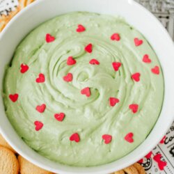 A bowl of creamy green Grinch-themed cookie dip topped with small red heart sprinkles, surrounded by cookies on a tray. The image has the text Grinch cookie dip at the top and homemadeheather.com at the bottom.