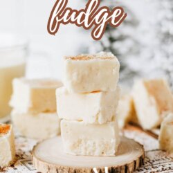 A stack of creamy eggnog fudge squares sits on a wooden slice, with more fudge pieces and a blurred festive background. Text reads Eggnog fudge—so good! homemadeheather.com.