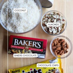 Top-down view of baking ingredients on a wooden surface, including a bowl of shredded coconut, a can of sweetened condensed milk, a bowl of sliced almonds, whole almonds, Bakerโs chocolate, and a bag of chocolate chips.