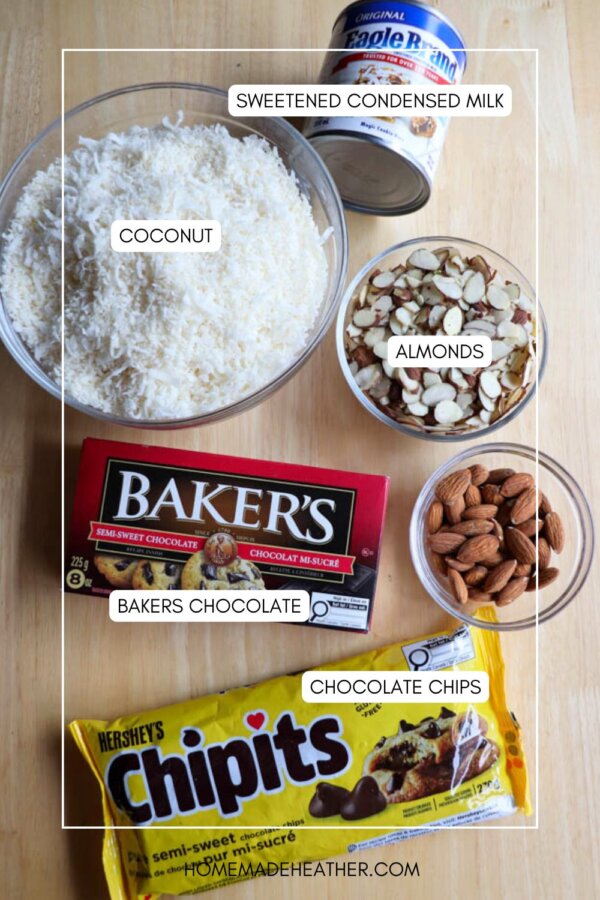 Top-down view of baking ingredients on a wooden surface, including a bowl of shredded coconut, a can of sweetened condensed milk, a bowl of sliced almonds, whole almonds, Bakerโs chocolate, and a bag of chocolate chips.