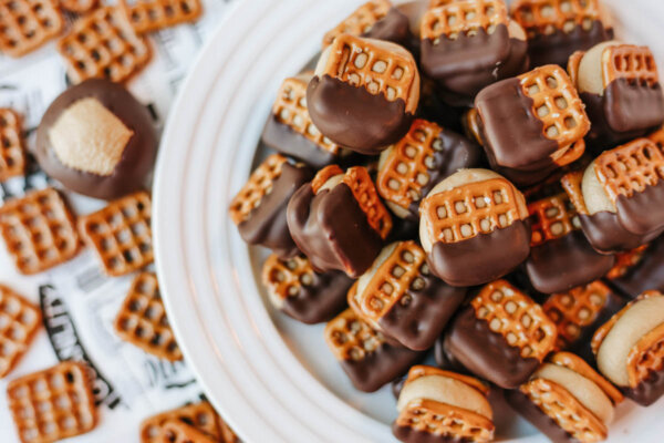 A white plate piled with pretzel sandwiches filled with peanut butter and half-dipped in chocolate, surrounded by more pretzel treats scattered on a surface.