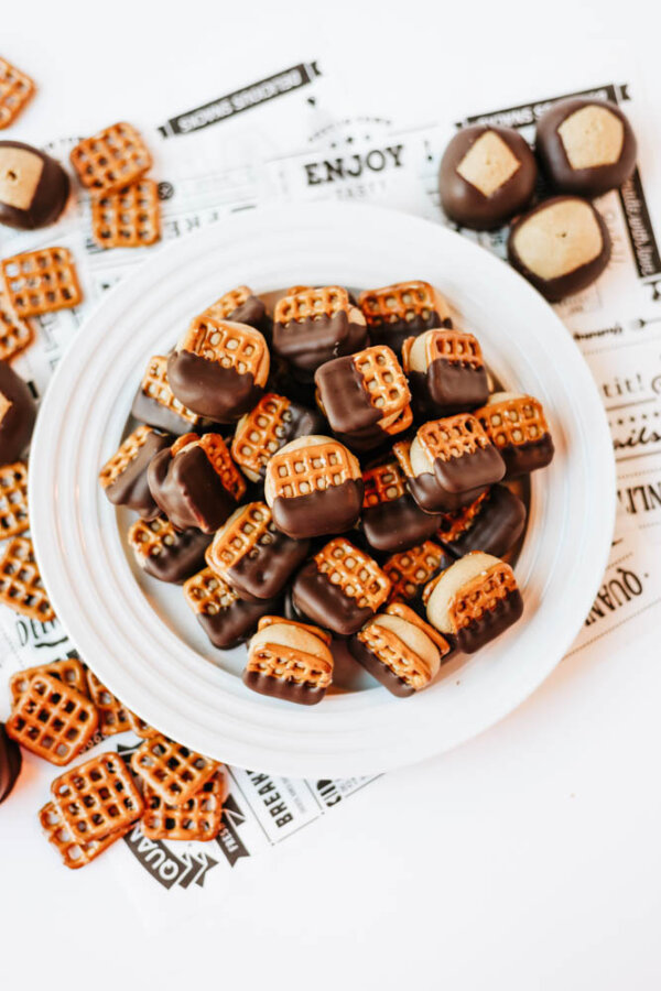 A white plate filled with square pretzels sandwiched with chocolate and peanut butter, surrounded by more pretzel sandwiches on a patterned paper background.