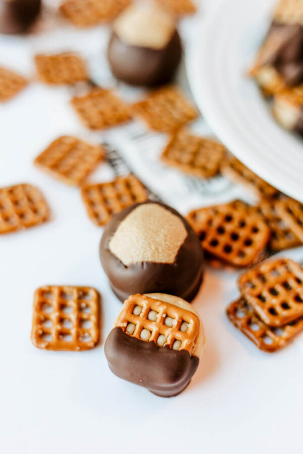 Chocolate-dipped pretzel squares and chocolate-covered peanut butter balls sprinkled with sugar are arranged on a white surface next to a white plate.