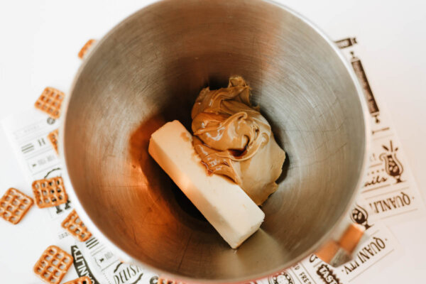 A metal mixing bowl containing a stick of butter and a scoop of peanut butter, with pretzels and printed paper partially visible underneath.