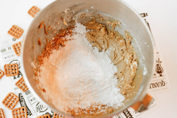 A metal mixing bowl with peanut butter dough and powdered sugar on top. Square pretzels are scattered nearby on a white surface with black-printed deli paper beneath the bowl.