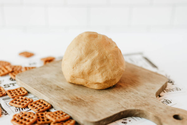 A ball of dough sits on a wooden cutting board, with small square pretzels scattered around on a white surface in the background.