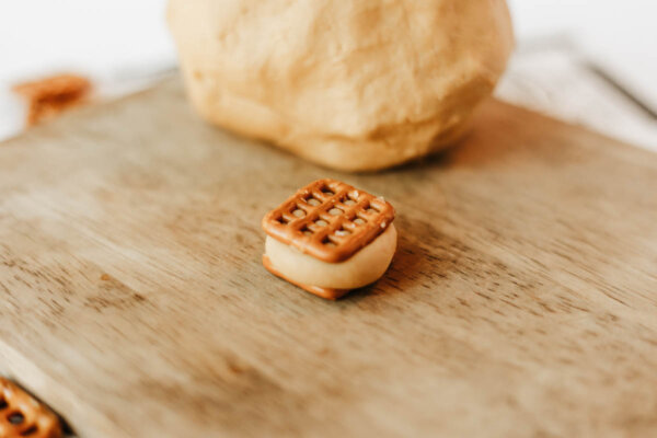 A small sandwich made of two square pretzels with peanut butter or dough in between sits on a wooden surface, with a larger lump of dough blurred in the background.