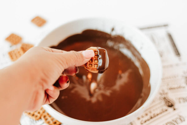A hand holding a small pretzel topped with caramel and dipped halfway into melted chocolate, with a bowl of melted chocolate and some pretzels in the background.