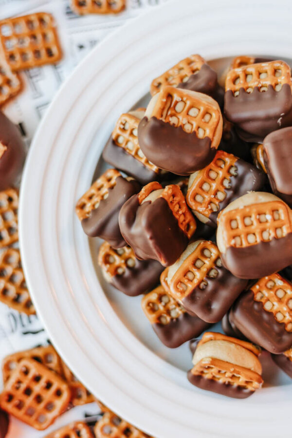 A plate filled with bite-sized pretzel sandwiches, each dipped halfway in chocolate. The sandwiches appear to have a peanut butter filling, with extra pretzels scattered around the plate.