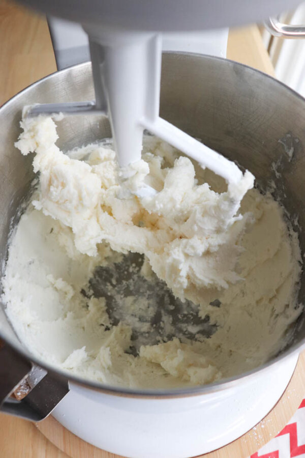 A metal mixing bowl with a stand mixer paddle attachment creaming butter and sugar, creating a fluffy, light mixture. The bowl sits on a wooden surface.