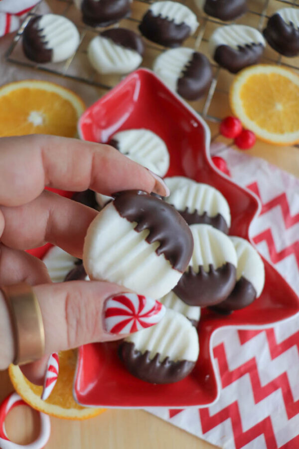 A hand with peppermint-themed nails holds a white, ridged cookie half-dipped in chocolate. More cookies are on a red, tree-shaped plate, with orange slices and a chevron napkin in the background.