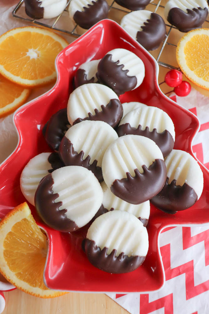 A red, tree-shaped plate holds several round, striped cookies dipped halfway in chocolate, surrounded by orange slices and holiday decorations. More cookies cool on a wire rack in the background.