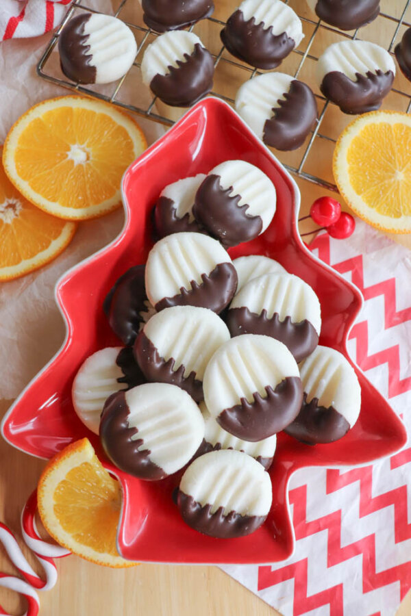 A red, tree-shaped plate holds white, round cookies half-dipped in chocolate. More cookies cool on a wire rack. Orange slices, red berries, candy canes, and a red chevron napkin decorate the scene.