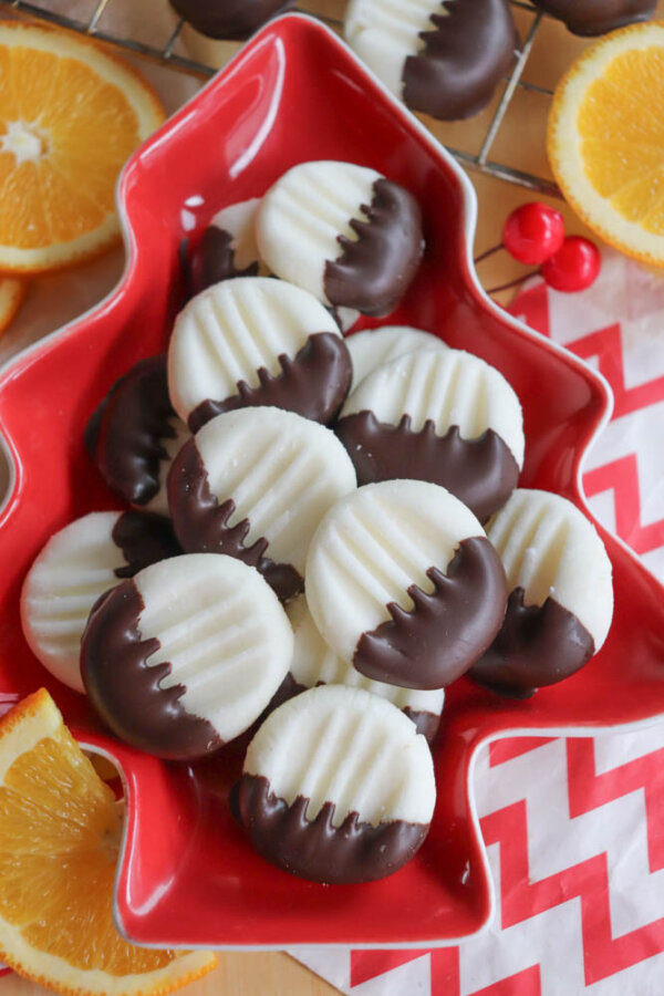Round white cookies partially dipped in chocolate are arranged on a red, tree-shaped plate. Orange slices and a red-and-white zigzag napkin are visible in the background.